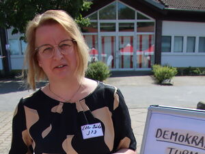 Draußen. Frau steht neben dem Jenga-Turm. Sie hat einen Holzklotz n beiden Händen. Darauf steht: Tierrechte. Die Frau ist blond, trägt ihr haare offen, sie trägt eine Brille.