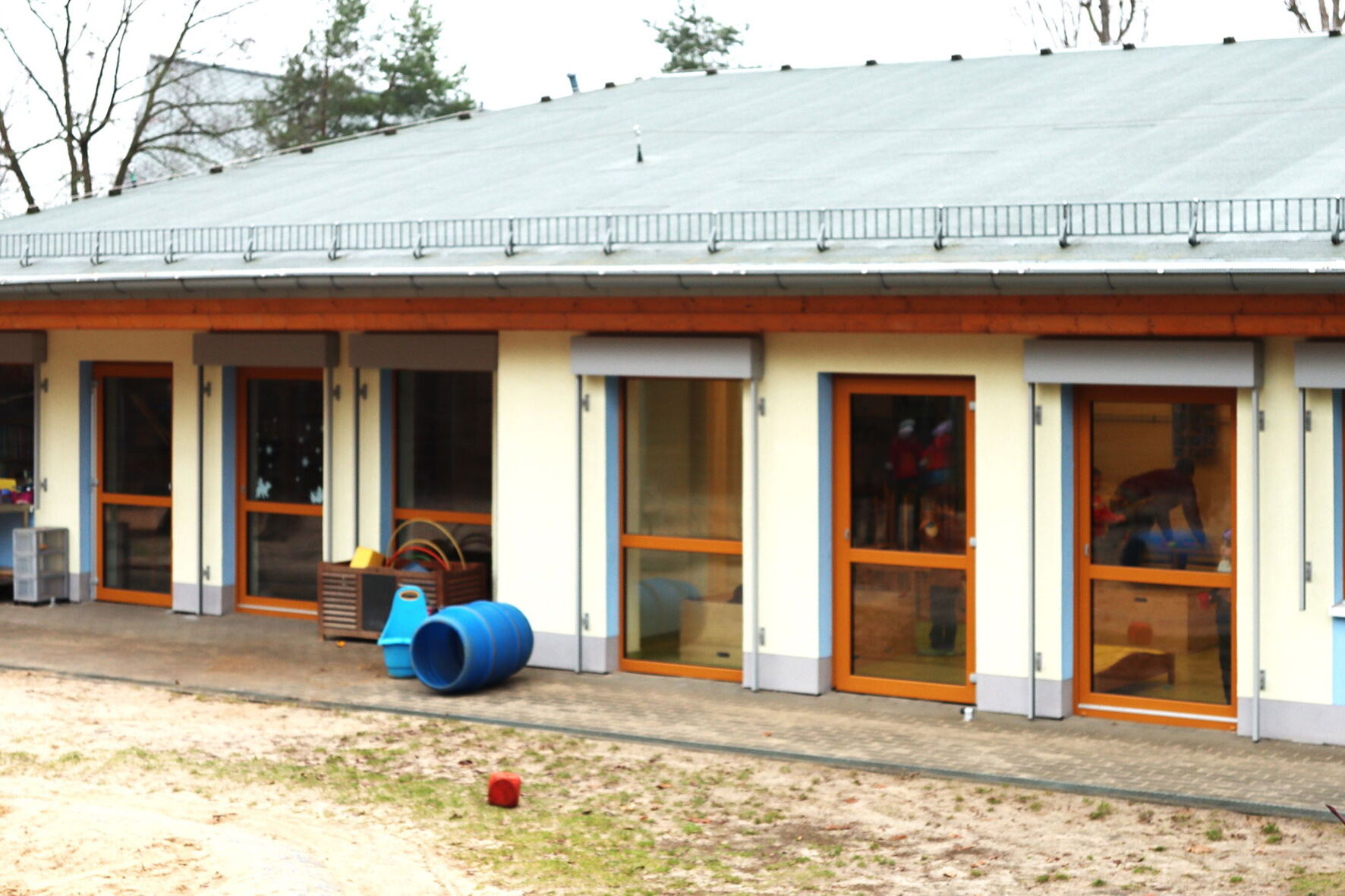 Draußen, kahle Bäume. Ein flaches Kita Gebäude mit vielen Fenstern und Terrassentüren.