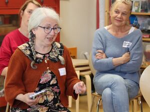 3 Personen sitzen auf Stühlen in einem Raum. Frau spricht und hält ein Notizbuch in der rechten Hand. Frau hat weiße Haare zu zwei Zöpfen geflochten und Brille. Frau schräg hinter ihr lächelt und sieht zu der Frau dir spricht.