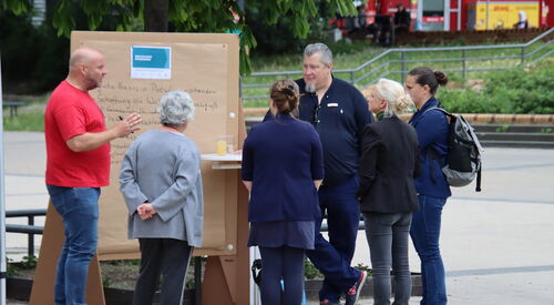 Draußen. 6 Personen stehen um einen Flipchart herum. 4 Politikerinnen unterhalten sich. 2 Männer stehen neben dem Flipshart. Einer im roten T-Shirt hält einen Stift in den Händen. Von weiterweg fotografiert.