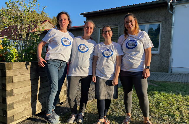 Draußen, Sonne scheint. 4 Frauen stehen nebeneinander und lächeln in die Kamera. Sie tragen einheitliche weiße T-Shirts mit Aufdruck: Blauer Kreis: Kinder stark machen steht drauf.