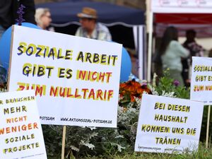 4 Protestplakate stecken in Blumenrabatte. Dahinter Pavillons und Menschen.