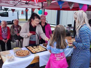 Am Kuchenstand gibt es reichlich Auswahl. Blechkuchen und andere Kuchen. Eine Frau gibt Kuchen an ein Mädchen.