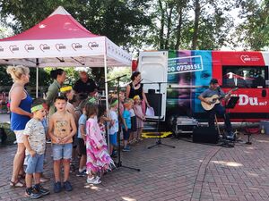 Draußen bei warmen Wetter. Ein roter AWO Pavillon und der AWO Ehrenamtsagentur Bus. Ein Mensch mit Gitarre und die Kinder stehen im Pavillon und singen. 3 Frauen stehen bei den Kindern.