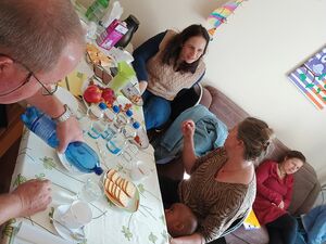 Gedeckter Tisch, mit Kaffee, Kuchen, Wasserflaschen. 3 Frauen, ein Kleinkind und ein Mann sitzen am Tisch. Zwei Frauen unterhalten sich.