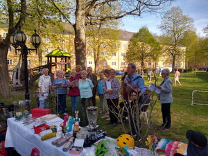 Der Chor bestehen aus 12 Menschen. Ein Mann mit Arkordion. Sie sttehen draußen im Schatten eines Baums.