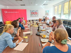 Großer Raum, große Tische. Viele Menschen sitzen sich gegenüber, einige machen sich Notizen.