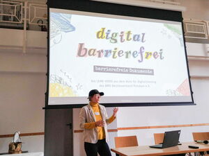 Auf einer Leinwand ist die Präsentation „digital barrierefrei – barrierefreie Dokumente“ zu sehen. Davor steht eine Frau mit Basecap und hält einen Vortrag.