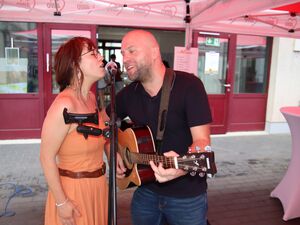 Frau und Mann singen in ein Mikro mit Ständer. Mann mit Gitarre. Die Frau hat braune lange Haare, Brille und trägt ein orangenes Kleid mit  Spaghettiträger, Mann mit Glatze, Bart, dunkelblaues Poloshirt und Jeans.