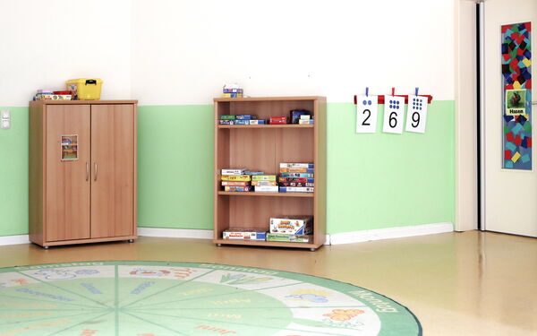 Spielzimmer. An der Wand ein offenes Regal mit Spielen. Auf dem Fußboben ein runder Kinderteppich mit den Wochentagen, und Monaten und dazu passenden Bildern.