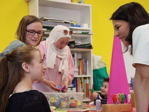 Ein Raum mit gelben Wänden. 3 Frauen, ein Mädchen, ein kleiner Junge. Frau mit dunklen schulterlangen Haaren lächelt Mädchen das zu ihr aufblickt. Eine pinke Zuckertüte steht auf dem Tisch.