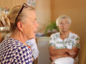 Nahaufnahme. Gesicht im Profil. Frau mit langen blonden Haaren, die zu einem Dutt am Hinterkopf zusammengebunden sind. Brille mit dunklem Rahmen ist auf den Kopf geschoben. Frau trägt kleine Ohrringe. Im Hintergrund unscharf ältere Frau.