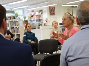 Zwei Rücken und Hinterköpfe von Männern, dazwischen hindurch zwei Frauen auf der gegenüberliegenden Seite. In der Mitte eine Frau mit blonden Haaren zum Dutt hochgesteckt, Brille auf den Kopf geschoben in einem rosa Oberteil, diskutiert lebhaft.