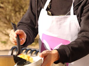 Draußen, Sonne scheint. Großküchen Topf eine Frau mit Schürze mit eine Keller befüllt eine Schale mit Essen.