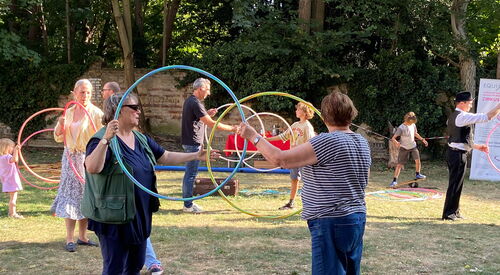 Jeweils zwei Personen halten zwei Hula Hoop Reifen senkrecht in den Händen. Mehrere Paare aus groß und klein stehen sich so gegenüber.