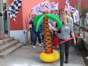 Eine Zielflagge links im Bild. Eine blonde Frau mit einer großen Gummi-Palme tritt über die Ziellinie (rotes Band) Hinter hier Teilnehmende im weißen AWO Flaggen.