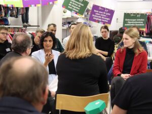 In einem Bekleidungs-Geschäft. Menschen von jung bis alt sitzen in zwei Stuhlkreisen. In der Mitte eine Frau mit dunklem lockigem Haar und weißem Hosenanzug, sie hat das Wort.