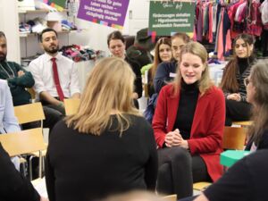 In einem Bekleidungs-Geschäft. Menschen von jung bis alt sitzen in zwei Stuhlkreisen. In der Mitte sitzen sich zwei Frauen gegenüber. Die Frau die spricht ist blond mit langen Haaren, schwarzer Kleidung unter einem roten Blazer.