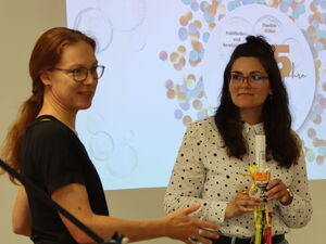 2 Frauen stehen im Veranstaltungsraum. Hinter ihnen die Präsentation.