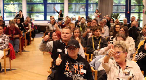 Voller Saal mit Teilnehmenden. Ein Mittelgang ist frei geräumt. Einige der Teilnehmenden lächeln und strecken den Daumen-Hoch.