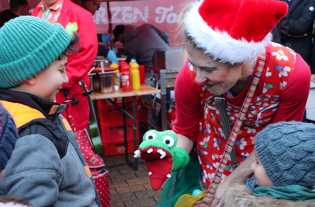 Clownin bückt sich zu zwei Kindern hinunter und lächelt es an. Sie hat eine Handpuppe als Krokodil, das reißt das Maul auf. Clownin hat eine rute Weihnachtmütze auf.