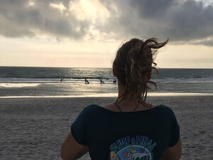 Strand, Meer, Sonne Wolken. Eine Frau mit dem Rücken zu Kamera blickt aufs Meer. Sie trägt ein "Surf and Heal" T-Shirt.