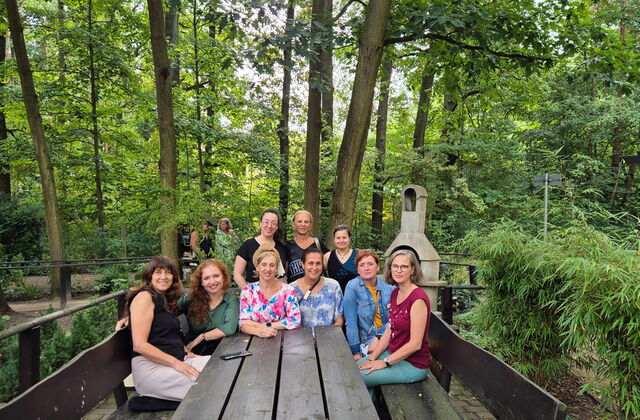 Gruppenfoto: 9 Personen sitzen im Halbrund an einem Holztisch, der in einem bewaldeten Gelände steht. Alle sehen in die Kamera.