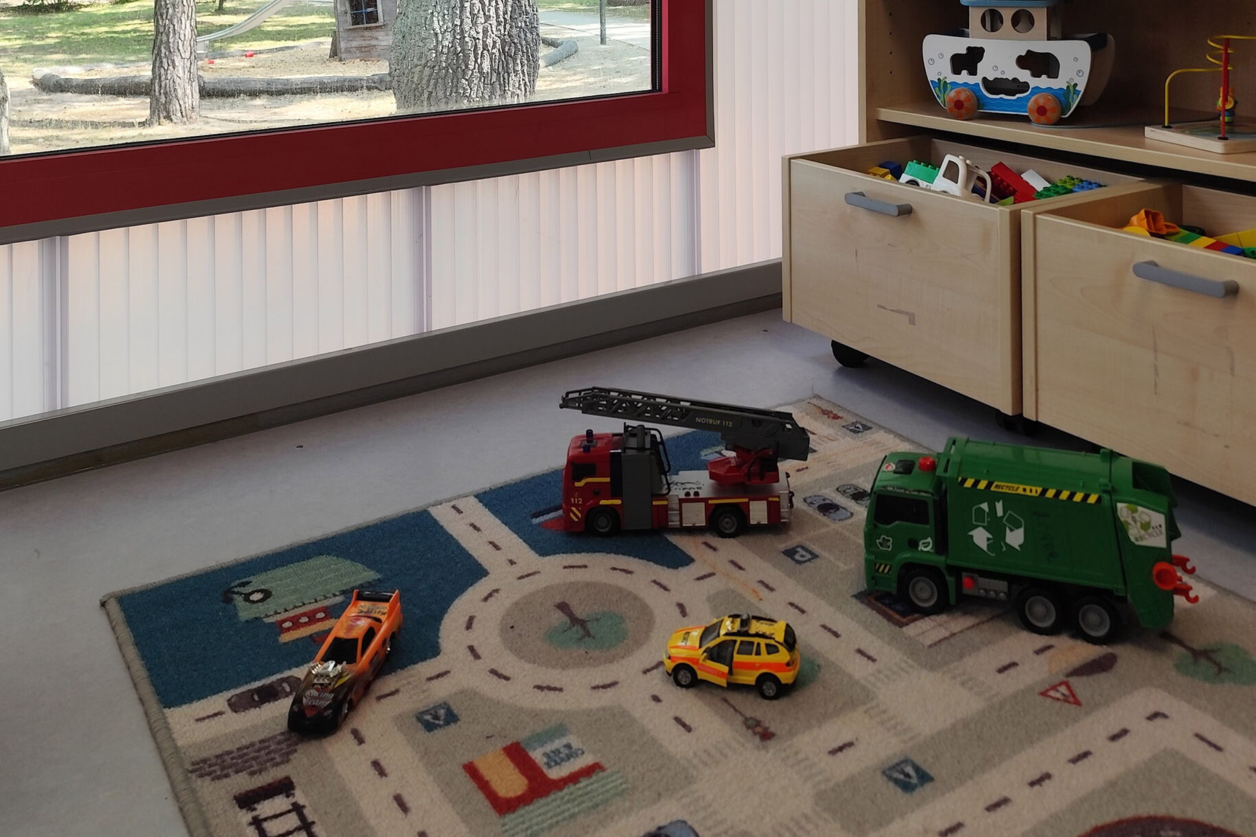 Teppich mit einem Straßenmuster, darauf sind Spielzeugautos und -figuren platziert. Im Hintergrund ist ein Holzregal mit Spielzeug und Pflanzen sowie ein Fenster, durch das Tageslicht hereinfällt.