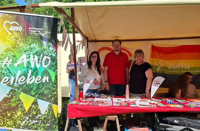 Drei Personen, zwei Frauen und ein Mann, stehen hinter einem Tisch in einem Marktstand.  Auf dem Tisch liegen Flyer, Stifte und Schlüsselbänder.
