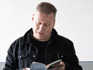 Innenraum. Mann in blauer Jeans, schwarzem Pullover  und Jacke, graue kurze Haare, liest interessiert in einem kleinen Buch / Broschüre. Eine Brille liegt neben ihm auf einem kleinen Tisch.