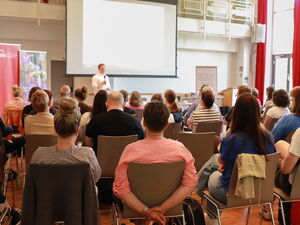 Saal. Teilnehmende von hinten. Vorn im Raum ein Mann mit Mikrofon.