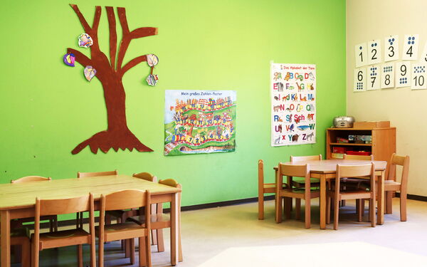 Zimmer mit kleinen Tischen und Stühlen. Eine Wand ist grün, daran hängen Poster mit Zahlen uns eins mit dem Alphabet.