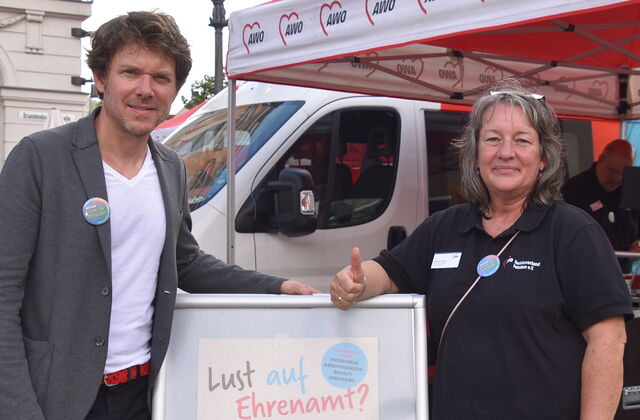 Draußen auf einem Platz. Ein Mann und eine Frau. Die Frau reckt dem Damen nach oben. Zwichen den Beiden ein Schild. Text: Lust auf Ehrenamt?