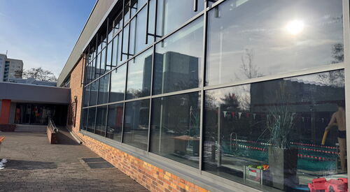 Schwimmhalle mit großem Fenster, ein großes Becken im inneren. Die Sonne spiegelt sich im Fenster.