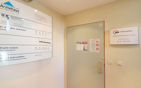 Flur. Geschlossene Tür zur Sozialstation. Rechts an der Wand ein großes Schild vom Ärztehaus worin sich die Sozialstation befindet.