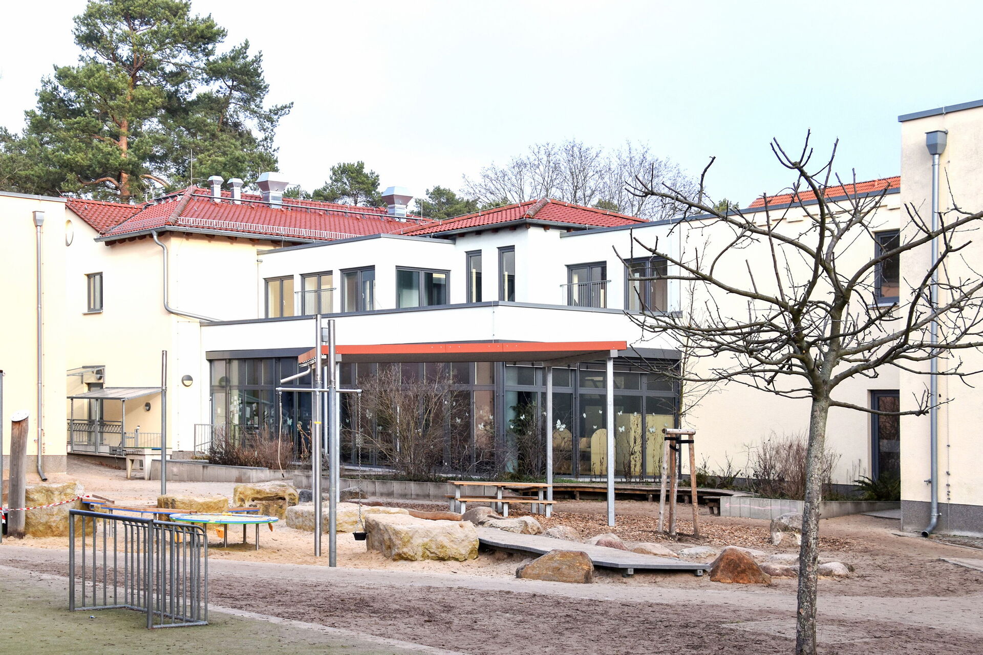 Zwei-stöckiges Kita Gebäude. Unten viele Fenster. Glas-Fassade. Davor kahle Bäumchen, ein Trampolin und Bänke.