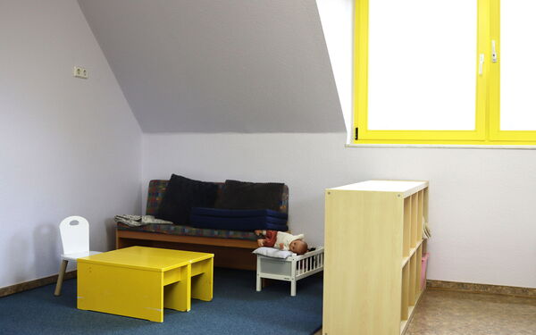 Spielzimmer. Blauer Teppich unter einer Dachschräge. An der Wand ein kleines Sofa mit Kissen, ein kleiner gelber Tisch steht davor, an dem Tisch ein kleiner weißer Stuhl. Neben dem Sofa ein  Puppenbett mit Babypuppe. Als Abgrenzung ein Regal mit Fächern.