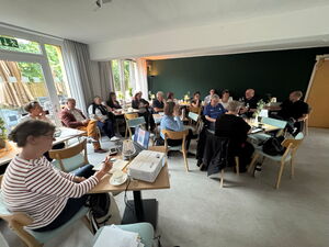 Sitzung: Größerer Raum, Menschen sitzen locker an Tischen.  Vor im Bild eine Frau an einem Tisch mit Laptop und Beamer.