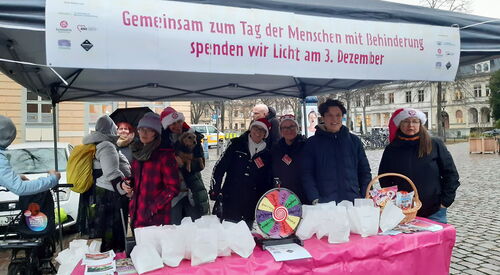 Pavillon, Tisch vorne auf dem kleine Papiertüten, kleines Glücksrad, ein Körbchen mit Süßigkeiten steht. Menschen stehen im Pavillon. Einige lächeln in die Kamera.