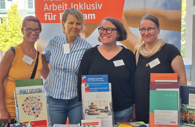 4 Frauen stehen nebeneinander am Stand des Netzwerk Arbeit inklusiv und lächeln in die Kamera. Vor ihnen auf dem Tisch Informationsmaterial zum Thema Arbeit mit Behinderung.
