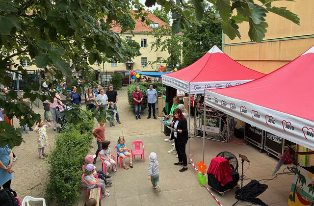 Draufsicht von weiterweg. Kita-Gelände. Rechts zwei rote AWO Pavillons, 4 Kinder sitzen auf kleinen pinken Stühlen. Erwachsene und weitere Kinder stehen herum.