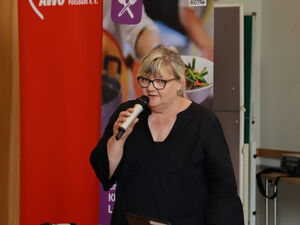 Frau mit Mikrofon. Frau trägt schwarz und steht vor einem lila Roll-Up der Initiative "Schule satt".