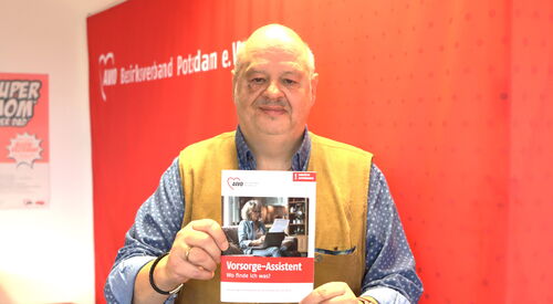 Portrait: Mann mit Glatze, geprunktem Hemd und gelber Weste darüber hält ein Heft / Broschüre vor sich.     Vorsorge-Assistent - Wo finde ich was?      steht darauf.