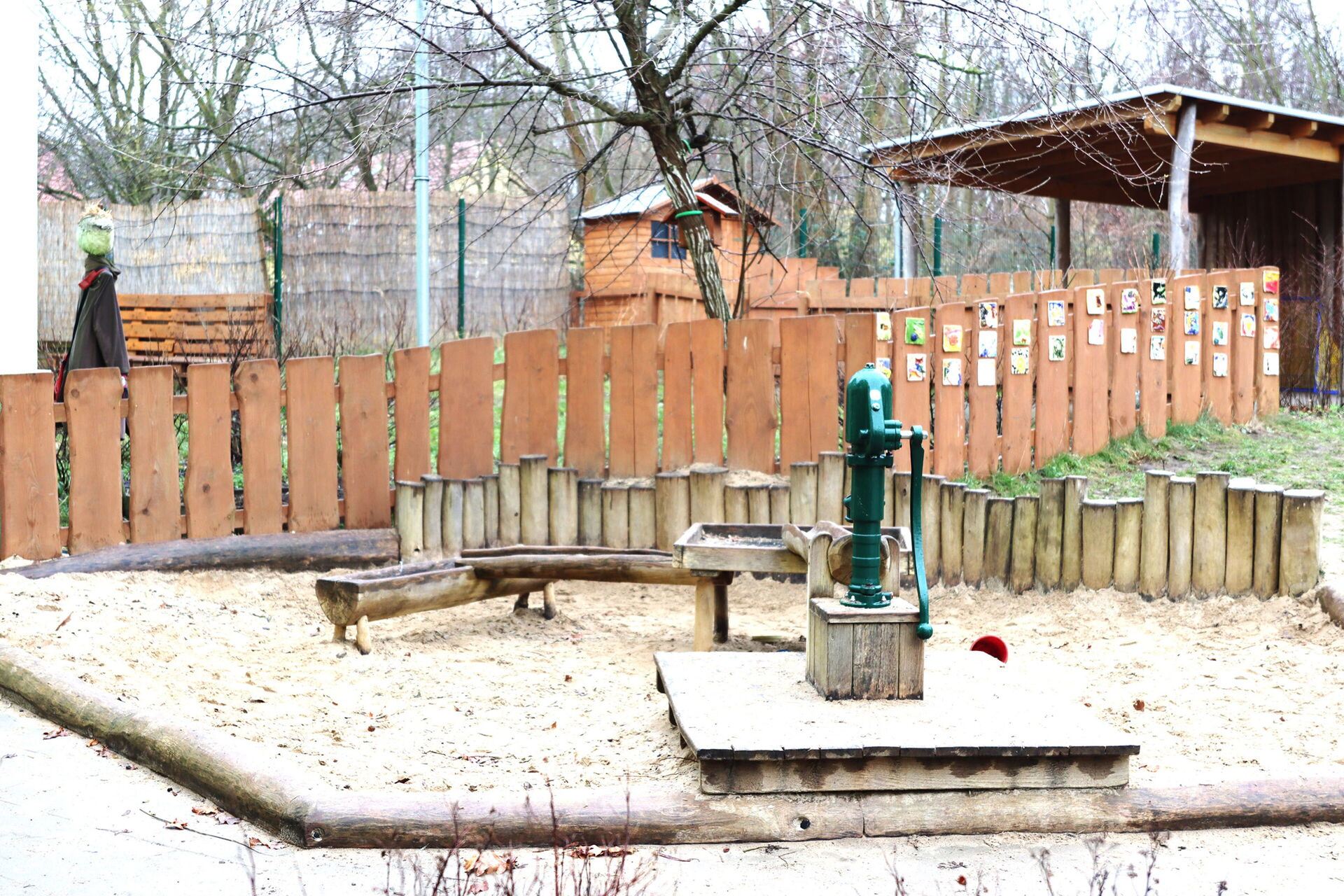 Draußen, kahle Bäume. Ein Wasserspielplatz. Sandkasten, eine Wasserpumpe mit Wasserbecken und zwei Wasserrinnen.