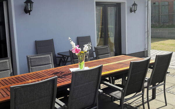 Große Terrasse mit langen Tisch und Stühlen vor einem Haus. Auf dem Tisch steht eine Vase mit Blumen. Die Sonne scheint.