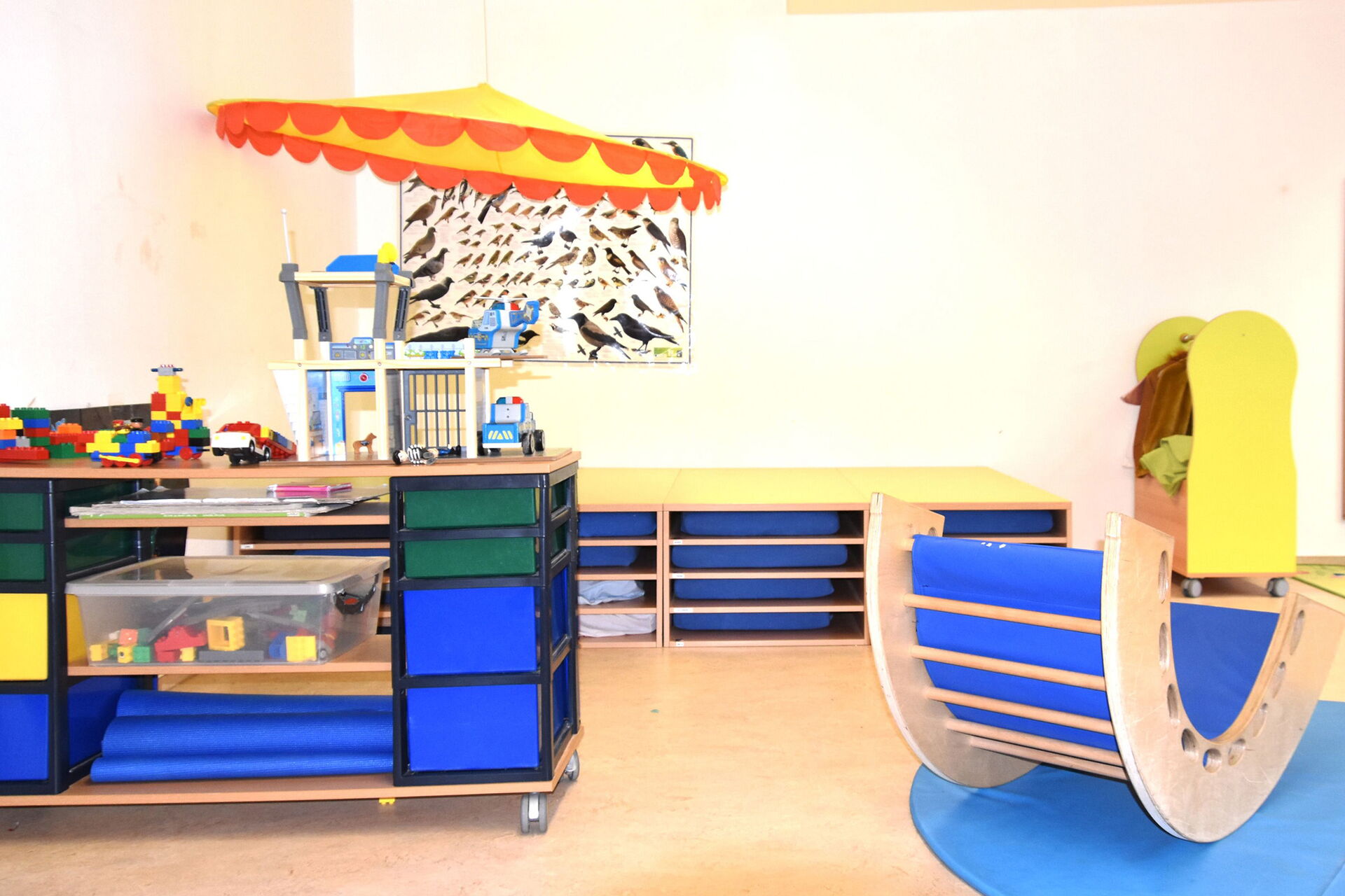 Spielzimmer. Kleines Regal mit Lego. Kleine halbrunde Wippe aus Holz mit blauer Polsterung innen. An der hinteren Wand ein Schaubild mit verschiedenen Vögeln.