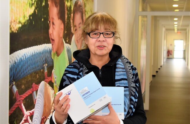 In einem Flur an der Wand ist ein Bild von spielenden Kindern. Frau mit brauen längeren Haaren, schwarzgerahmte Brille, Kette und offener dunklen Jacke hält Kartons mit Corona Tests in beinen Händen. Frau lächelt in die Kamera.