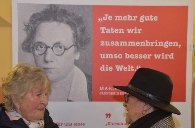 Hintergrund: Plakat mit schwarz-weiß Foto von Marie Juchacz mit Zitat: Je mehr gute Taten wir zusammenbringen, umso besser wird die Welt. Davor zwei ältere Frauen im Gespräch.