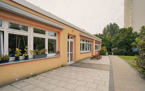 Flaches langes Gebäude mit orangener Fassade. Auf dem Fensterbrett stehen Pflanzen.