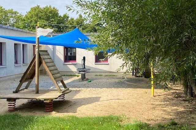 Kleiner Spielplatz vor einem Gebäude. Ein blaues Sonnensegel ist darüber gespannt und sorgt für Schatten.
